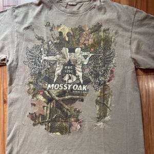 Op de afbeelding: Een beige T-shirt met een camouflageprint met een afbeelding van een jager met pijl en boog. De afbeelding bevat ook de tekst "EST. 1986" en "MOSSY OAK BRAND CAMO".
