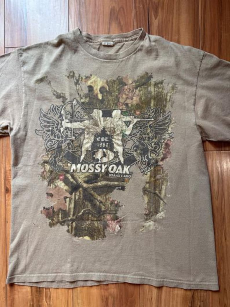 Op de afbeelding: Een beige T-shirt met een camouflageprint met een afbeelding van een jager met pijl en boog. De afbeelding bevat ook de tekst "EST. 1986" en "MOSSY OAK BRAND CAMO".