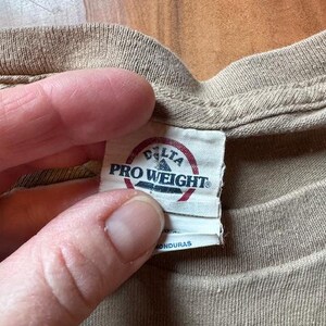 Op de afbeelding: Een beige T-shirt met een wit label waarop "Delta Pro Weight" en "Gemaakt in Honduras" staat.