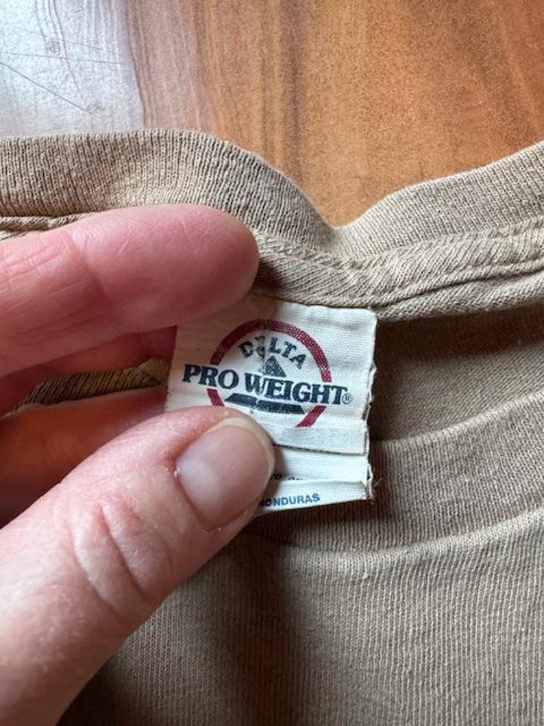 Op de afbeelding: Een beige T-shirt met een wit label waarop "Delta Pro Weight" en "Gemaakt in Honduras" staat.