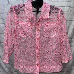 May include: A pink button-down shirt with a floral pattern. The sheer blouse has two chest pockets and three-quarter sleeves. The shirt is displayed on a black hanger against a white brick wall.