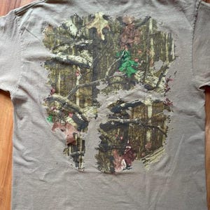 Op de afbeelding: Een bruin T-shirt met een camouflagepatroon gedrukt op de achterkant. Het patroon heeft tinten groen, bruin en beige.