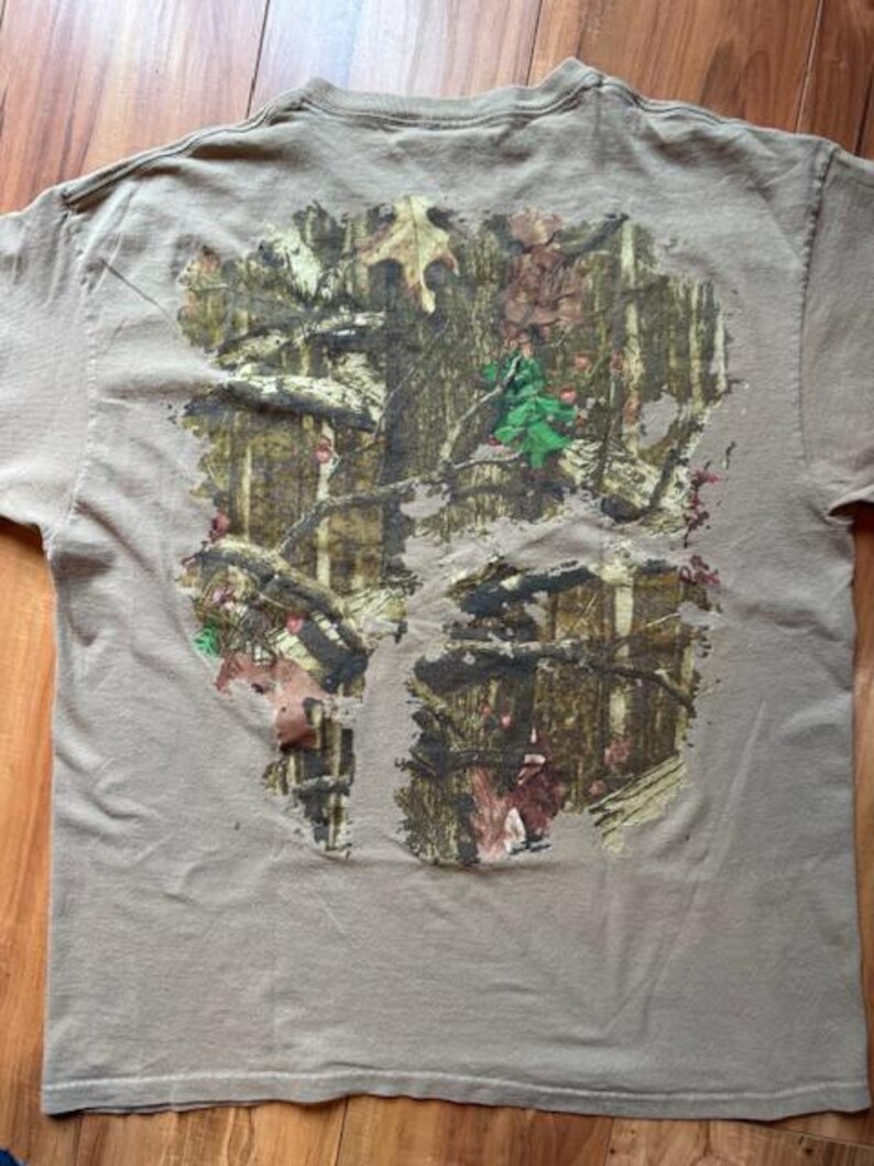 Op de afbeelding: Een bruin T-shirt met een camouflagepatroon gedrukt op de achterkant. Het patroon heeft tinten groen, bruin en beige.