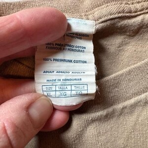 Op de afbeelding: Een wit label met zwarte tekst op een bruin t-shirt. Het label zegt "100% voorgekrompen katoen" en "Adult Adulto Adulte". Het label heeft ook een maattabel met de maten XL, XG en TO.