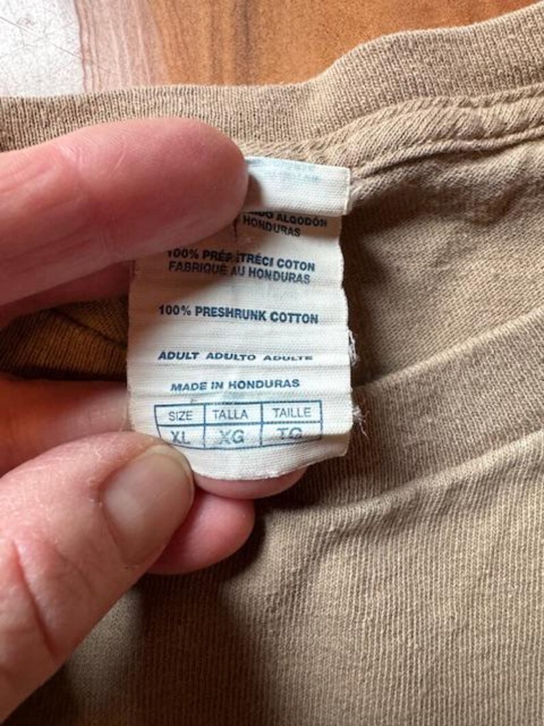 Op de afbeelding: Een wit label met zwarte tekst op een bruin t-shirt. Het label zegt "100% voorgekrompen katoen" en "Adult Adulto Adulte". Het label heeft ook een maattabel met de maten XL, XG en TO.