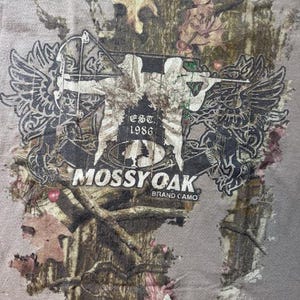 Op de afbeelding: Een bruin T-shirt met een camouflagepatroon met een afbeelding van een persoon met een boog en pijl, twee vleugels en de tekst "Mossy Oak Brand Camo" en "Est. 1986".