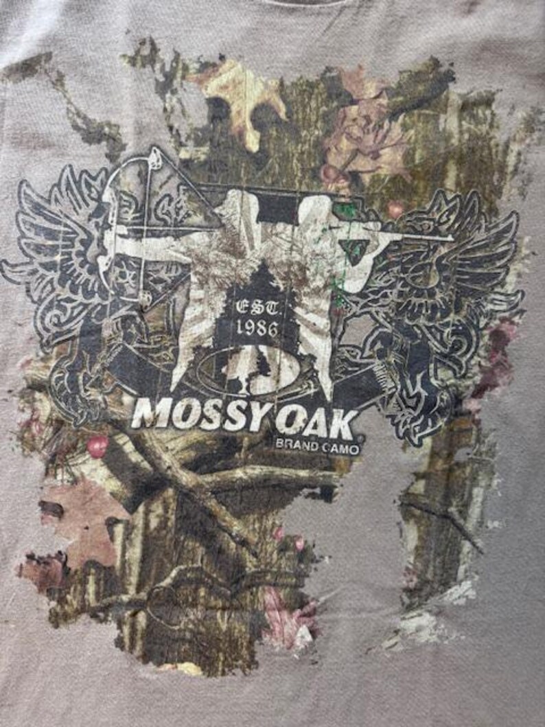 Op de afbeelding: Een bruin T-shirt met een camouflagepatroon met een afbeelding van een persoon met een boog en pijl, twee vleugels en de tekst "Mossy Oak Brand Camo" en "Est. 1986".