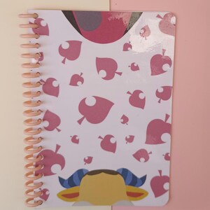 Könnte beinhalten: Ein spiralgebundenes Notizbuch mit einem rosa-weißen Blättermuster. Das Notizbuch hat eine pfirsichfarbene Spiralbindung. Der untere Teil des Notizbuchs zeigt eine Cartoonfigur in Gelb, Blau und Braun.