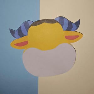 Peut inclure: Découpage en papier représentant une tête de taureau de dessin animé. Le taureau a une fourrure jaune, des oreilles intérieures roses et des cornes rayées bleues et noires. La tête est posée sur un fond de papier bleu et beige.