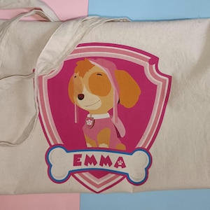 May include: A cream tote bag with a pink and white shield design featuring a cartoon dog character in a pink outfit and hood. The name "EMMA" is printed in white on a bone-shaped design. The bag has a handle.