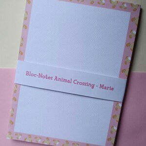Peut inclure: Un bloc-notes avec une bordure rose ornée d'un motif répété de personnages de dessins animés. Le bloc-notes porte une bande blanche avec le texte "Bloc-Notes Animal Crossing - Marie". Le fond est rose clair.