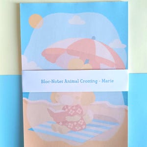 May include: A notepad featuring a beach scene with a sun, clouds, and an umbrella. The notepad has a light blue background with the text "Bloc-Notes Animal Crossing - Marie" on a white band. The bottom of the notepad shows a character reading a book.