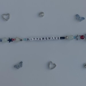 Pu&ograve; includere: Un braccialetto di perline con la parola "BITTERSUITE" scritta con perline bianche. Il braccialetto presenta varie perline blu, rosse e trasparenti, insieme a ciondoli a forma di stella e farfalla. Ciondoli a forma di cuore e spirale argentati sono sparsi.