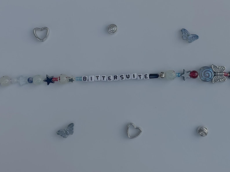 Pu&ograve; includere: Un braccialetto di perline con la parola "BITTERSUITE" scritta con perline bianche. Il braccialetto presenta varie perline blu, rosse e trasparenti, insieme a ciondoli a forma di stella e farfalla. Ciondoli a forma di cuore e spirale argentati sono sparsi.