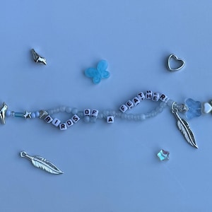Pu&ograve; includere: Un braccialetto di perline azzurre con la scritta "BIRDS OF A FEATHER". Il braccialetto presenta vari ciondoli, tra cui una piuma d'argento, un uccello, un cuore e una farfalla blu. Le perline sono azzurre e bianche.