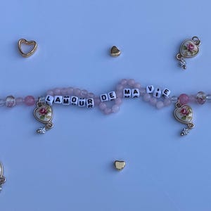 Pu&ograve; includere: Una collana di perline con perline e ciondoli rosa. La collana presenta ciondoli a forma di cuore con disegni floreali e la frase "AMOUR DE MA VIE" scritta con perline di lettere bianche. Accenti dorati a forma di cuore sono sparsi.