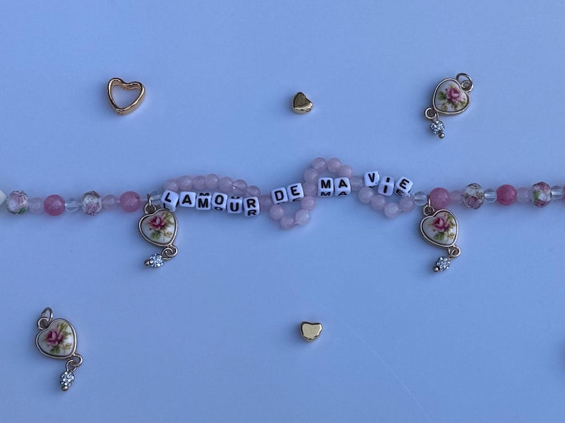 Pu&ograve; includere: Una collana di perline con perline e ciondoli rosa. La collana presenta ciondoli a forma di cuore con disegni floreali e la frase "AMOUR DE MA VIE" scritta con perline di lettere bianche. Accenti dorati a forma di cuore sono sparsi.