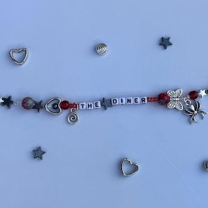 Pu&ograve; includere: Un braccialetto di perline con la scritta "THE DINER" in perline bianche. Il braccialetto presenta perline rosse, grigie e argentate, ciondoli a forma di stella e cuore e un ciondolo a forma di farfalla e ragno. Ulteriori ciondoli argentati e forme di stella sono sparsi.