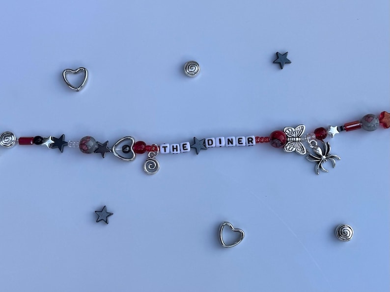 Pu&ograve; includere: Un braccialetto di perline con la scritta "THE DINER" in perline bianche. Il braccialetto presenta perline rosse, grigie e argentate, ciondoli a forma di stella e cuore e un ciondolo a forma di farfalla e ragno. Ulteriori ciondoli argentati e forme di stella sono sparsi.