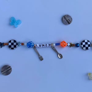 Pu&ograve; includere: Una collana di perline con la parola "LUNCH" scritta con perline bianche. La collana presenta varie perline blu, arancioni, bianche e nere e ciondoli argentati a forma di forchetta e cucchiaio. Sono inclusi anche accenti decorativi a farfalla e a spirale.