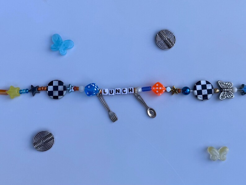 Pu&ograve; includere: Una collana di perline con la parola "LUNCH" scritta con perline bianche. La collana presenta varie perline blu, arancioni, bianche e nere e ciondoli argentati a forma di forchetta e cucchiaio. Sono inclusi anche accenti decorativi a farfalla e a spirale.