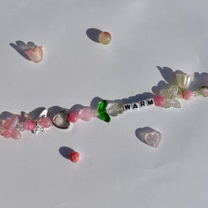 K&ouml;nnte beinhalten: Eine Halskette mit Perlen aus rosa und klaren Perlen, silbernen Schmetterlings- und Herzanh&auml;ngern und dem Wort "WARM" in wei&szlig;en Buchstabenperlen. Die Halskette hat blumen- und herzf&ouml;rmige Perlen.