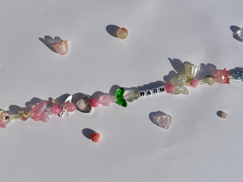 K&ouml;nnte beinhalten: Eine Halskette mit Perlen aus rosa und klaren Perlen, silbernen Schmetterlings- und Herzanh&auml;ngern und dem Wort "WARM" in wei&szlig;en Buchstabenperlen. Die Halskette hat blumen- und herzf&ouml;rmige Perlen.