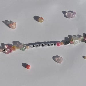 K&ouml;nnte beinhalten: Ein Armband mit Perlen, auf dem der Name "HAMPSTEAD" mit wei&szlig;en Buchstabenperlen geschrieben steht. Das Armband hat rosa, klare und silberne Perlen sowie Schmetterlings- und Herzanh&auml;nger. Zus&auml;tzliche dekorative Perlen und Anh&auml;nger sind verstreut.