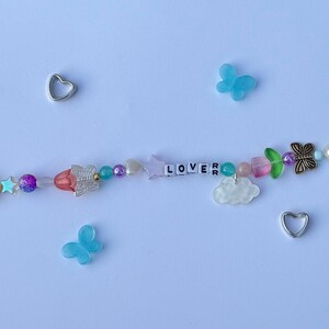以下が含まれることがあります： 白い文字ビーズで「LOVER」と書かれたビーズブレスレット。ブレスレットには、ピンク、青、紫のビーズ、蝶と星のチャームが付いています。2つのシルバーのハートチャームと2つの青い蝶のチャームが散らばっています。