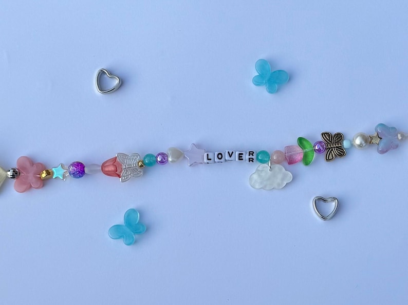 以下が含まれることがあります： 白い文字ビーズで「LOVER」と書かれたビーズブレスレット。ブレスレットには、ピンク、青、紫のビーズ、蝶と星のチャームが付いています。2つのシルバーのハートチャームと2つの青い蝶のチャームが散らばっています。