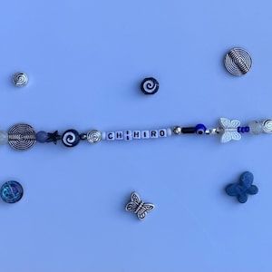 Pu&ograve; includere: Un braccialetto di perline con il nome "CHIRO" scritto con perline di lettere bianche. Il braccialetto presenta perline blu, argento e bianche, insieme a ciondoli a forma di farfalla e stella. Sono presenti anche ciondoli a spirale e farfalla argentati.
