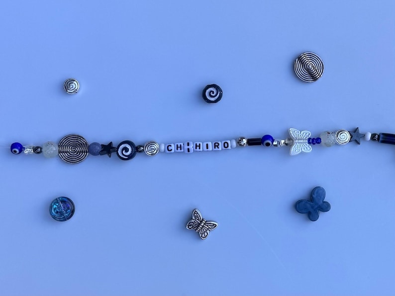 Pu&ograve; includere: Un braccialetto di perline con il nome "CHIRO" scritto con perline di lettere bianche. Il braccialetto presenta perline blu, argento e bianche, insieme a ciondoli a forma di farfalla e stella. Sono presenti anche ciondoli a spirale e farfalla argentati.