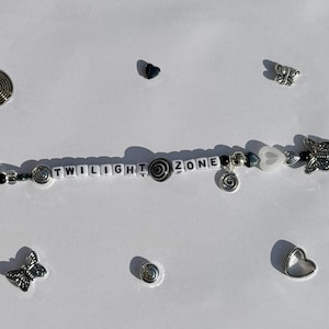 K&ouml;nnte beinhalten: Ein Armband mit Perlen, auf dem die Worte "TWILIGHT ZONE" mit wei&szlig;en Buchstabenperlen geschrieben sind. Es hat silberfarbene Spiral-, Herz- und Schmetterlingsanh&auml;nger sowie schwarze und wei&szlig;e Perlen. Zus&auml;tzliche Anh&auml;nger und Verzierungen sind verstreut.