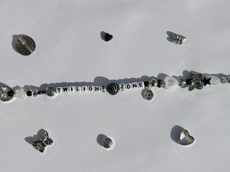 K&ouml;nnte beinhalten: Ein Armband mit Perlen, auf dem die Worte "TWILIGHT ZONE" mit wei&szlig;en Buchstabenperlen geschrieben sind. Es hat silberfarbene Spiral-, Herz- und Schmetterlingsanh&auml;nger sowie schwarze und wei&szlig;e Perlen. Zus&auml;tzliche Anh&auml;nger und Verzierungen sind verstreut.