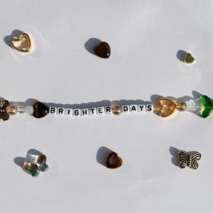 K&ouml;nnte beinhalten: Ein Perlenarmband mit den Worten "BRIGHTER DAYS" in wei&szlig;en Buchstabenperlen. Das Armband hat verschiedene Charms, darunter goldene Herzen, Schmetterlinge und gr&uuml;ne blattf&ouml;rmige Perlen. Zus&auml;tzliche Herz- und Schmetterlings-Charms sind verstreut.