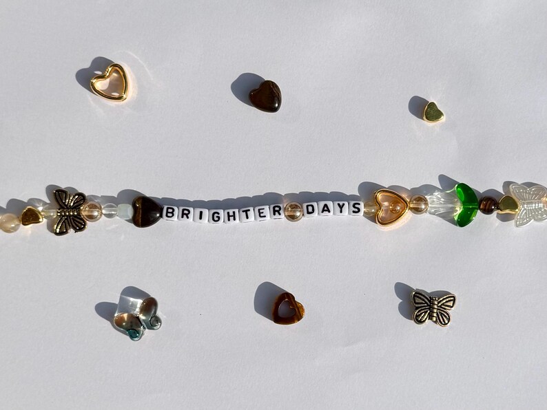 K&ouml;nnte beinhalten: Ein Perlenarmband mit den Worten "BRIGHTER DAYS" in wei&szlig;en Buchstabenperlen. Das Armband hat verschiedene Charms, darunter goldene Herzen, Schmetterlinge und gr&uuml;ne blattf&ouml;rmige Perlen. Zus&auml;tzliche Herz- und Schmetterlings-Charms sind verstreut.