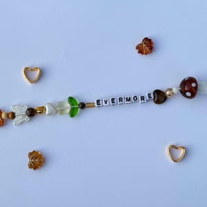 以下が含まれることがあります： 白い文字ビーズで「EVERMORE」と書かれたビーズブレスレット。ブレスレットには、葉や蝶のチャームなど、茶色、金色、緑色のさまざまなビーズが使用されています。2つの金のハートチャームと葉の形をしたビーズが散らばっています。