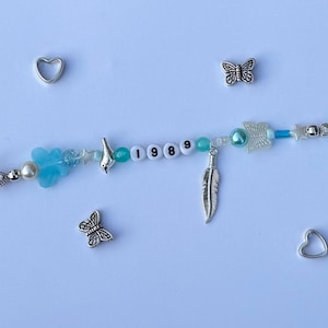 以下が含まれることがあります： 青、白、シルバーのアクセントが付いたビーズブレスレット。ブレスレットには、ビーズ、チャーム、白い文字ビーズで綴られた数字「1989」が含まれています。チャームには、蝶、ハート、羽、鳥が含まれています。