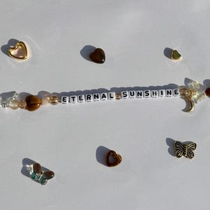 K&ouml;nnte beinhalten: Ein Perlenaccessoire mit dem Text "ETERNAL SUNSHINE" in wei&szlig;en Buchstabenperlen. Das Accessoire verf&uuml;gt &uuml;ber verschiedene Charms, darunter Schmetterlinge, Herzen und einen Halbmond. Die Farbpalette umfasst goldene, braune und transparente Perlen.