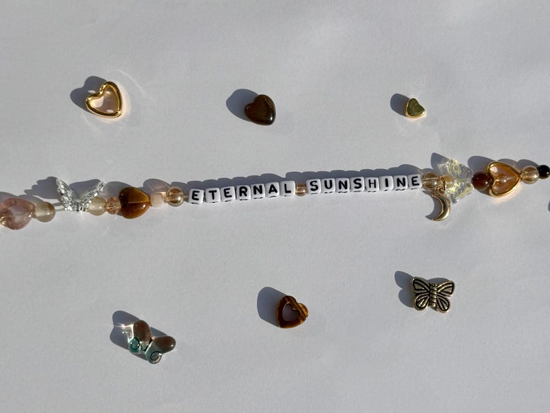 K&ouml;nnte beinhalten: Ein Perlenaccessoire mit dem Text "ETERNAL SUNSHINE" in wei&szlig;en Buchstabenperlen. Das Accessoire verf&uuml;gt &uuml;ber verschiedene Charms, darunter Schmetterlinge, Herzen und einen Halbmond. Die Farbpalette umfasst goldene, braune und transparente Perlen.