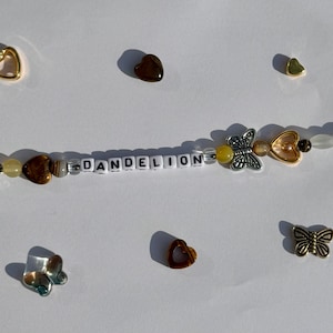 K&ouml;nnte beinhalten: Eine Perlenkette mit dem Wort "DANDELION" aus wei&szlig;en Buchstabenperlen. Die Kette hat verschiedene Perlen in Braun-, Gelb- und Klart&ouml;nen sowie Schmetterlings- und herzf&ouml;rmige Anh&auml;nger. Zus&auml;tzliche Herz- und Schmetterlingsanh&auml;nger sind verstreut.