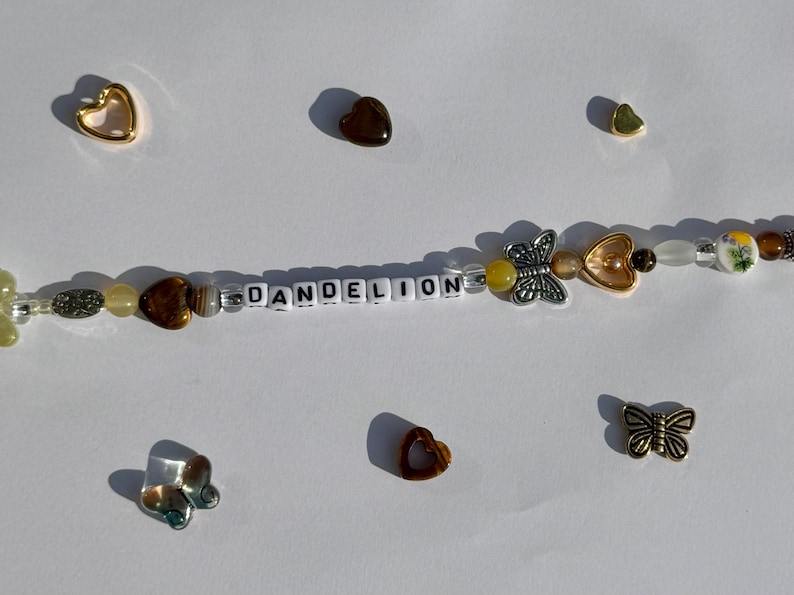 K&ouml;nnte beinhalten: Eine Perlenkette mit dem Wort "DANDELION" aus wei&szlig;en Buchstabenperlen. Die Kette hat verschiedene Perlen in Braun-, Gelb- und Klart&ouml;nen sowie Schmetterlings- und herzf&ouml;rmige Anh&auml;nger. Zus&auml;tzliche Herz- und Schmetterlingsanh&auml;nger sind verstreut.