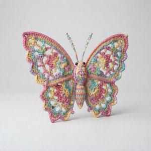 Könnte beinhalten: Ein gehäkelter Schmetterling mit filigranen, durchbrochenen Flügeln in Pastelltönen wie Rosa, Gelb, Blau und Lavendel. Der Körper ist in passenden Farben gestreift und die Fühler sind mit kleinen, durchscheinenden, bunten Perlen verziert.