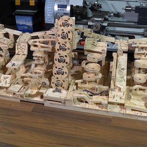 Könnte beinhalten: Eine komplexe kinetische Holzskulptur mit Zahnrädern, Rampen und Bahnen. Die hellfarbene Holzkonstruktion umfasst mehrere Ebenen und komplizierte mechanische Elemente. Die Skulptur ist für die Bewegung von Objekten konzipiert.