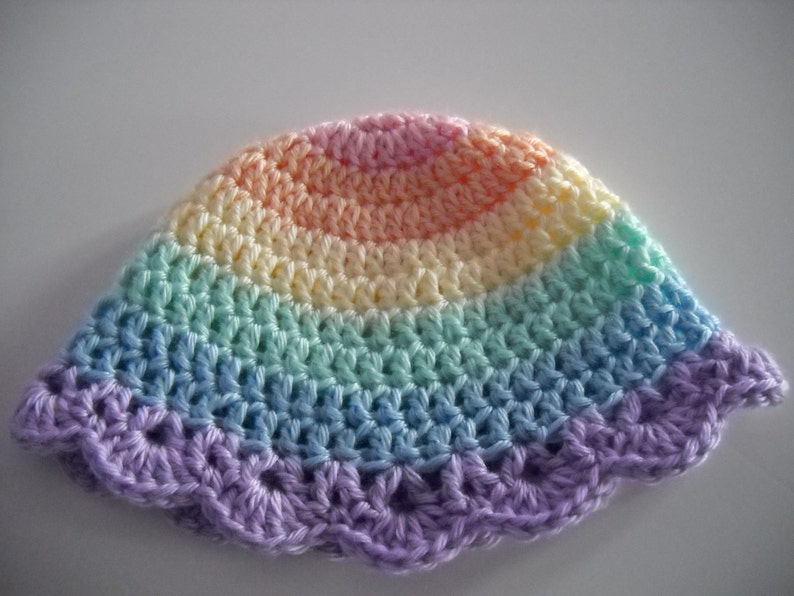 Puede incluir: Un gorro de ganchillo con un patr&oacute;n de degradado de arco&iacute;ris en tonos pastel. El dise&ntilde;o transita desde un rosa suave en la coronilla pasando por amarillo, verde menta y azul claro, con un borde festoneado morado en la parte inferior.