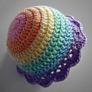 Puede incluir: Un gorro de ganchillo redondo con un degradado de colores pastel que incluye rosa, naranja, amarillo, verde y azul, terminado con un borde festoneado de color morado. La textura del hilo es visible en toda la estructura abovedada.