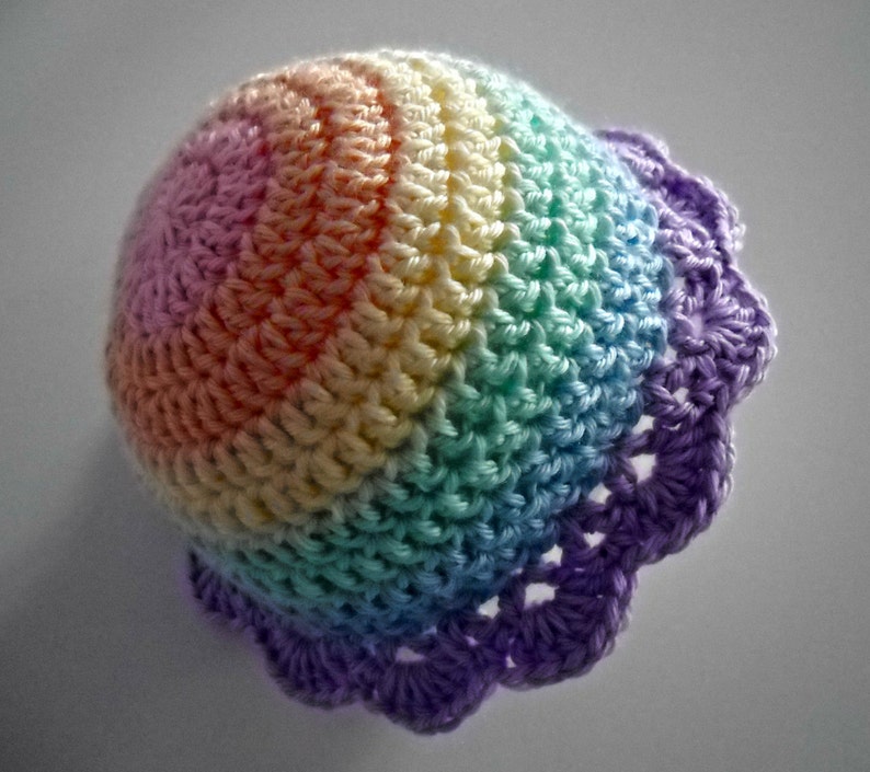 Puede incluir: Un gorro de ganchillo redondo con un degradado de colores pastel que incluye rosa, naranja, amarillo, verde y azul, terminado con un borde festoneado de color morado. La textura del hilo es visible en toda la estructura abovedada.