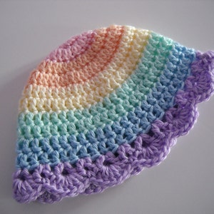 Puede incluir: Un gorro de ganchillo con un patr&oacute;n de arco&iacute;ris que presenta bandas conc&eacute;ntricas de rosa pastel, amarillo, verde menta y azul claro, terminado con un borde festoneado de color morado. El art&iacute;culo tiene una textura de punto visible.