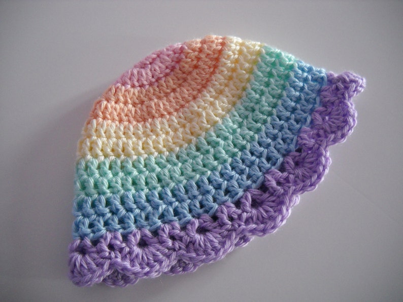 Puede incluir: Un gorro de ganchillo con un patr&oacute;n de arco&iacute;ris que presenta bandas conc&eacute;ntricas de rosa pastel, amarillo, verde menta y azul claro, terminado con un borde festoneado de color morado. El art&iacute;culo tiene una textura de punto visible.