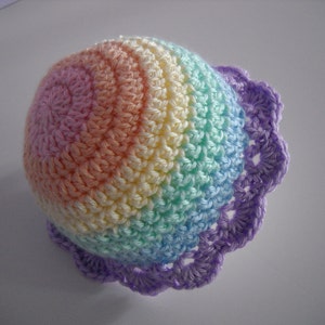 Puede incluir: Un gorro de ganchillo con un patr&oacute;n de degradado de arco&iacute;ris en tonos pastel de hilo rosa, naranja, amarillo, verde y azul. El gorro est&aacute; rematado con un borde festoneado de color morado, creando un dise&ntilde;o circular texturizado con puntos visibles.